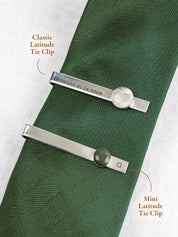 Two tie clips on a green tie with text indicating the size of the round stone set on the clip, the larger 'Classic Latitude Tie Clip' and the smaller 'Mini Latitude Tie Clip'