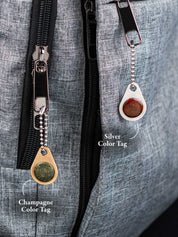 Two metal zipper pull tags set with natural stone, one silver color and one champagne color, attached to a gray backpack