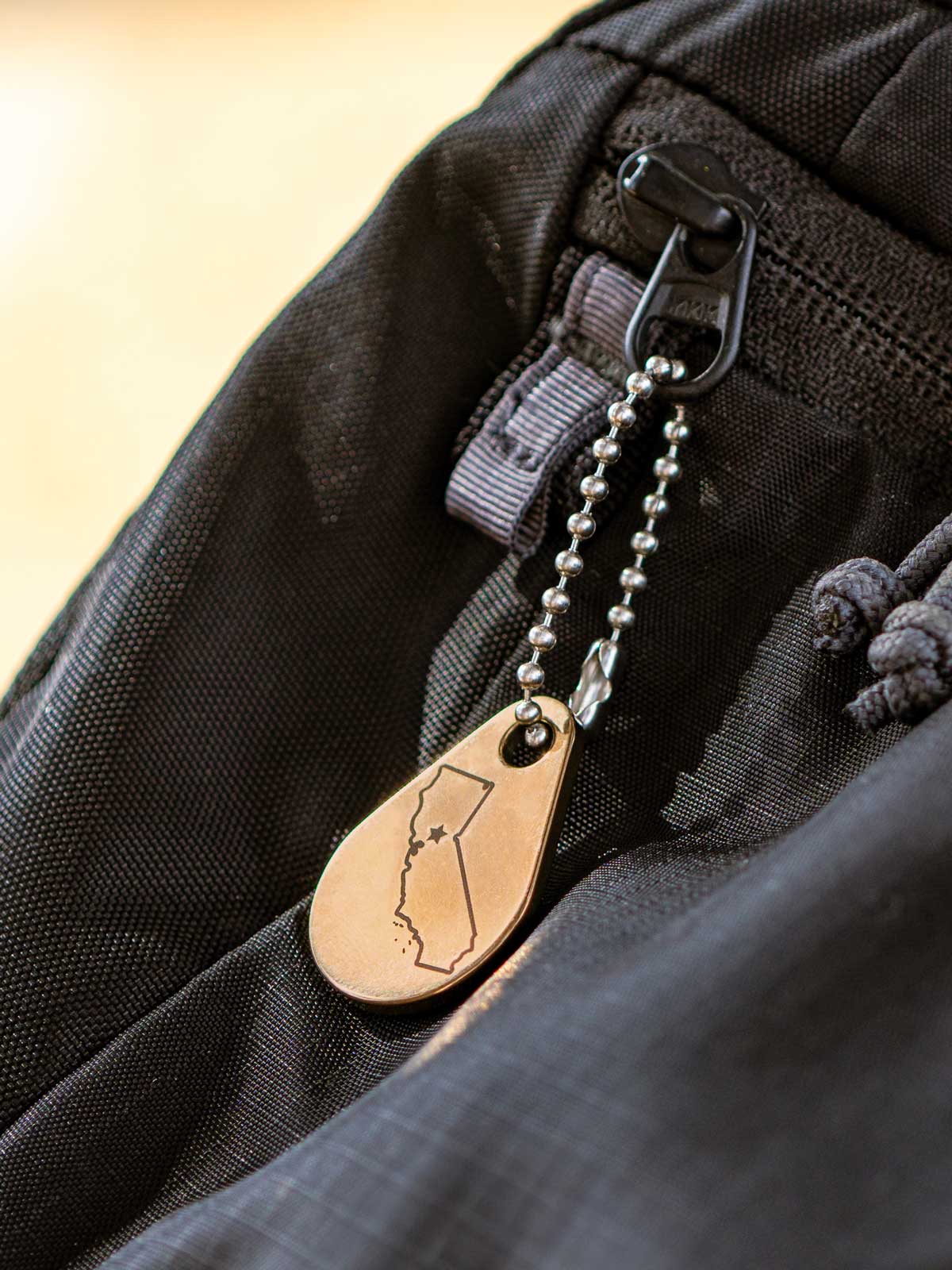 Ball chain custom zipper pull with a metal tag personalized with an engraving of the state of California and attached to a black backpack