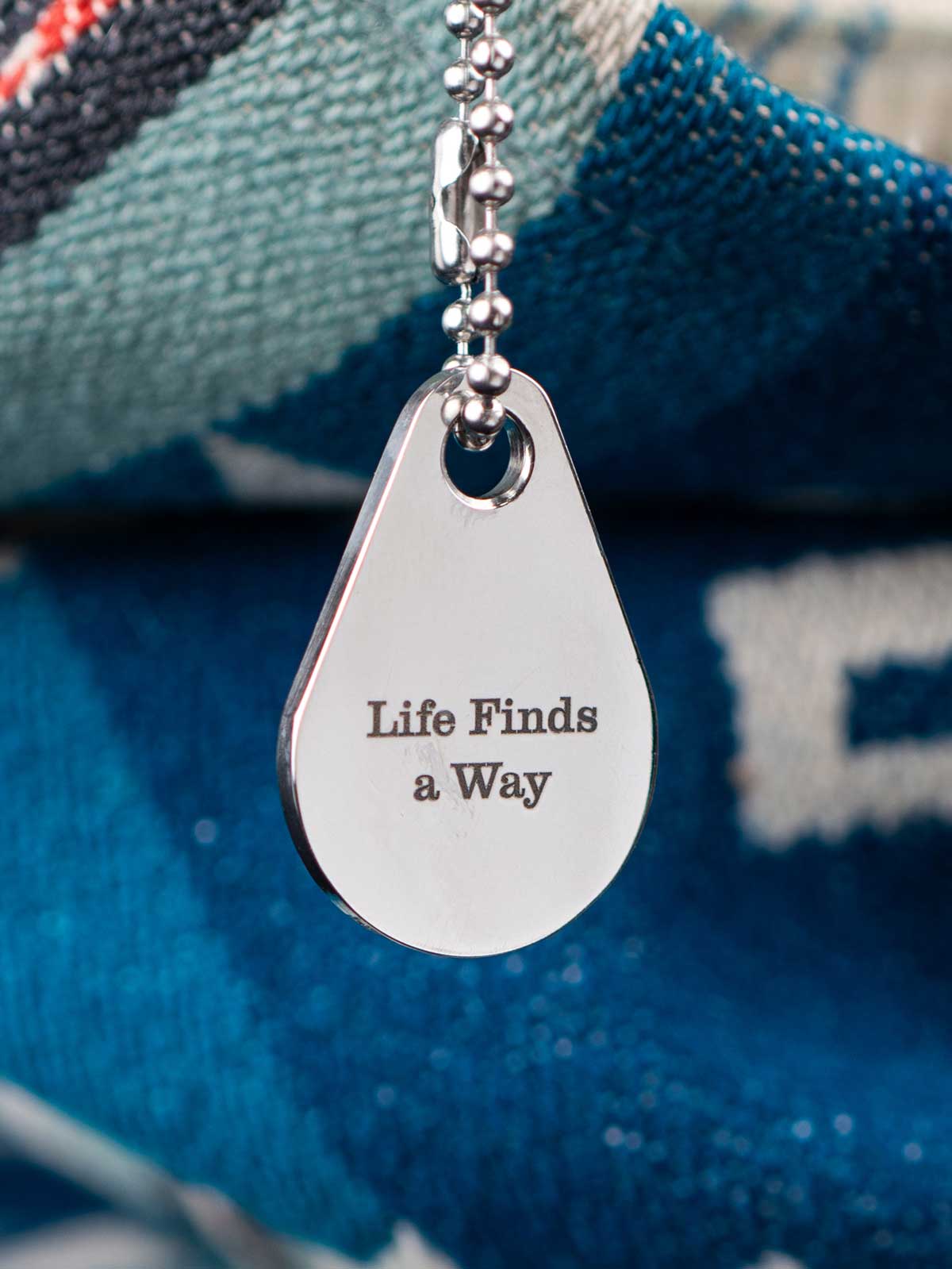 Teardrop-shaped metal zipper pull with 'Life Finds a Way' engraving on a blue bag