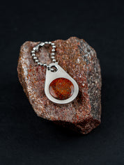 Teardrop-shaped zipper pull on a ball chain set with a red stone and sitting on the customer's original rock it was made from