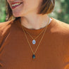 A woman in a burnt orange t-shirt smiles while wearing layered gold necklaces