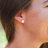 A woman wears white round stud earrings made with natural stone. The stone is set in a 14k gold cup.