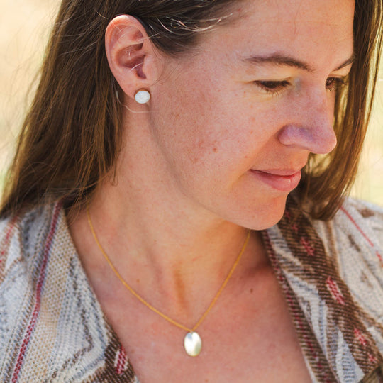 A woman in a boho-style jacket wears a pair of round stone stud earrings set in 14k gold, paired with a gold fill charm necklace