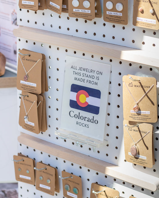 A market display holds a variety of stone jewelry along with a sign that reads, "All jewelry on this stand is made from Colorado rocks"