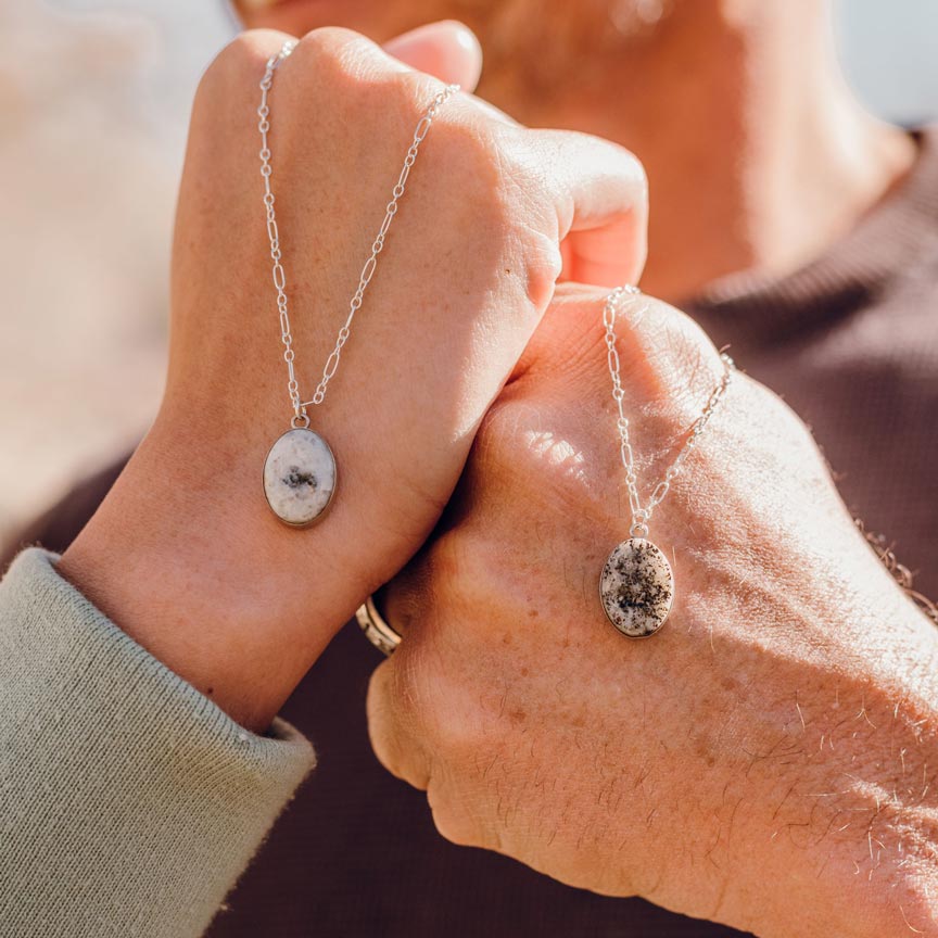 A couple clasps hands while holding matching white stone pendant necklaces