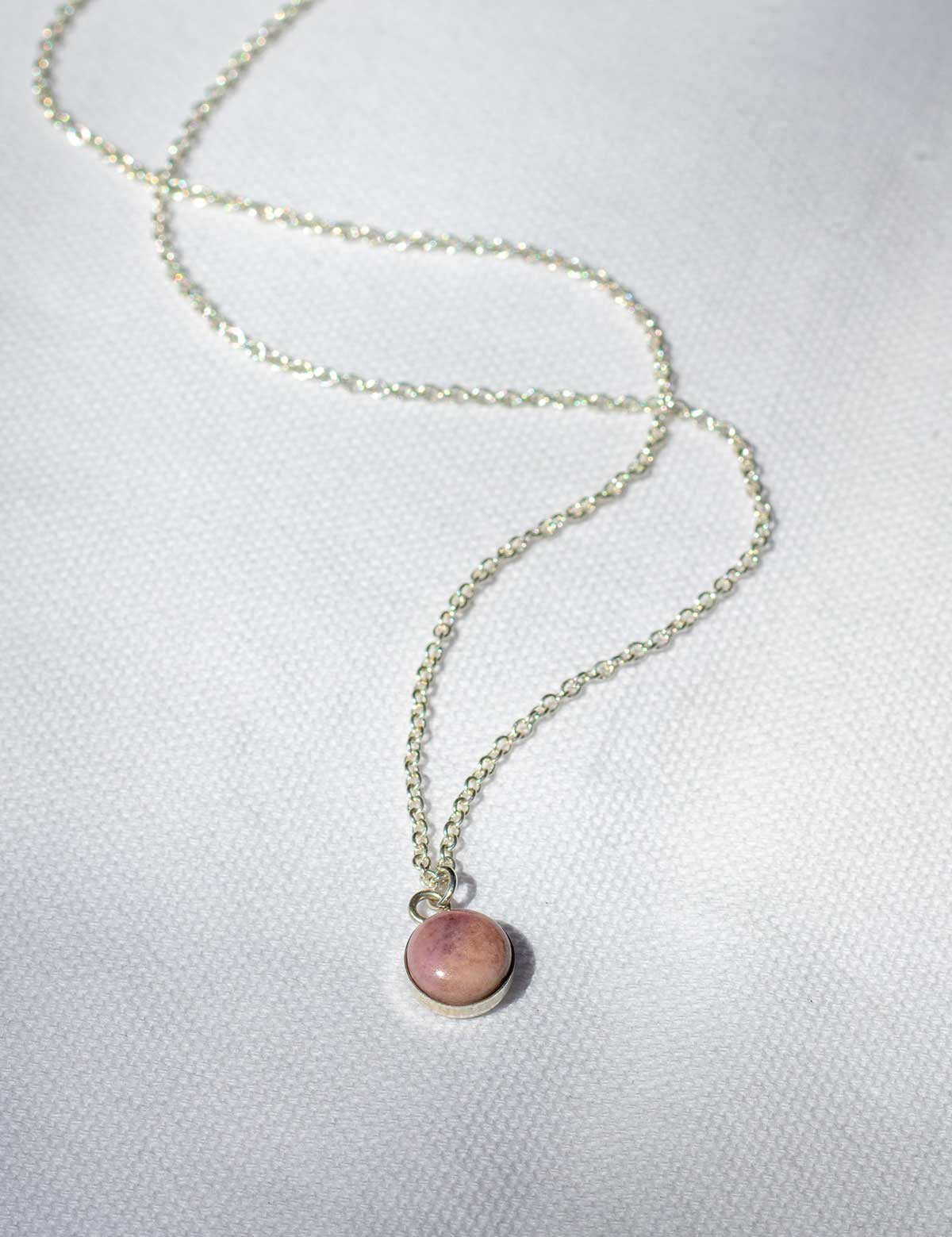 A natural pink rock is set in sterling silver to make a small, round charm that hangs from a necklace chain.