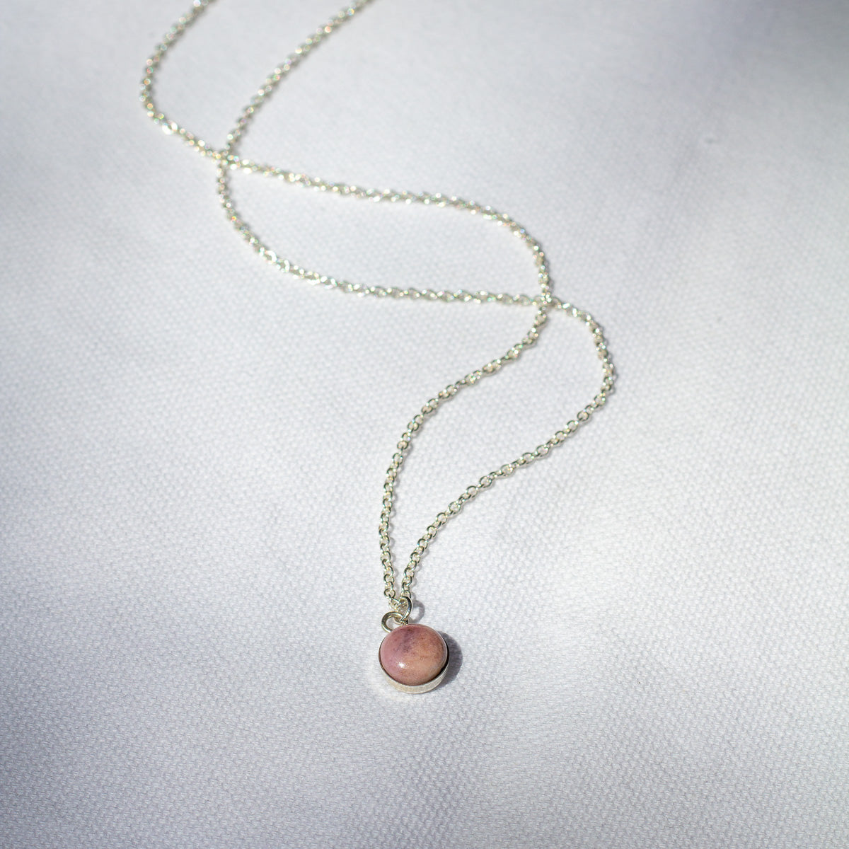A natural pink rock is set in sterling silver to make a small, round charm that hangs from a necklace chain.