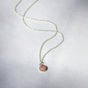 A natural pink rock is set in sterling silver to make a small, round charm that hangs from a necklace chain.